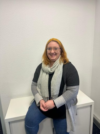 Lächelnde Frau mit blonden Haaren und Schal sitzt auf einer weißen Box in einem hellen Raum.
