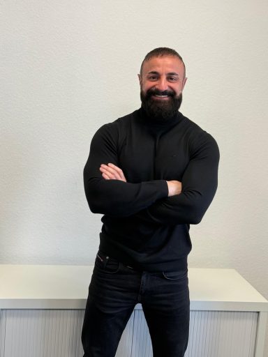 Mann mit Bart und muskulösem Oberkörper, in schwarzem Outfit, lächelt freundlich.