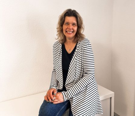 Frau mit lockigem Haar sitzt auf einer Bank, trägt gestreifte Jacke und lächelt freundlich.