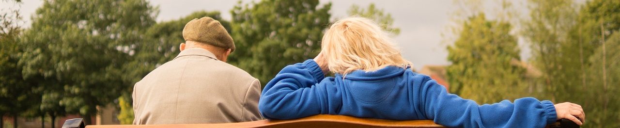 Zwei Personen sitzen auf einer Bank, Rücken zu Kamera, umgeben von Bäumen.