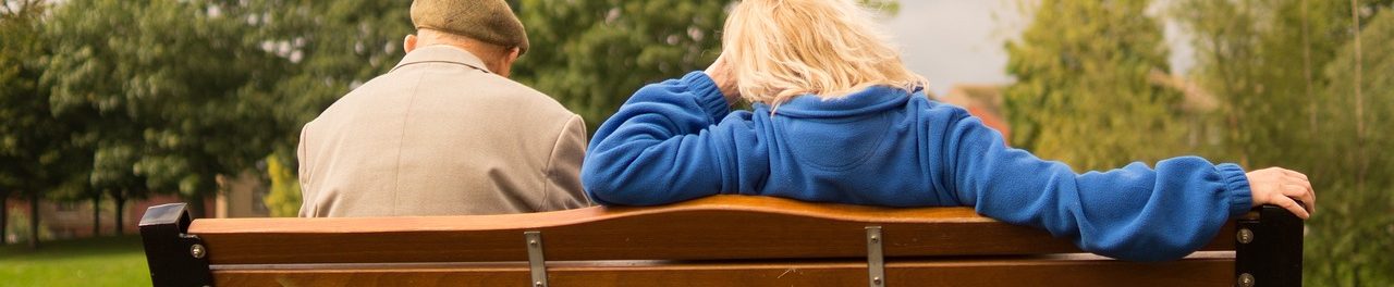 Paar sitzt auf einer Parkbank, Rückenansicht, umgeben von Bäumen.