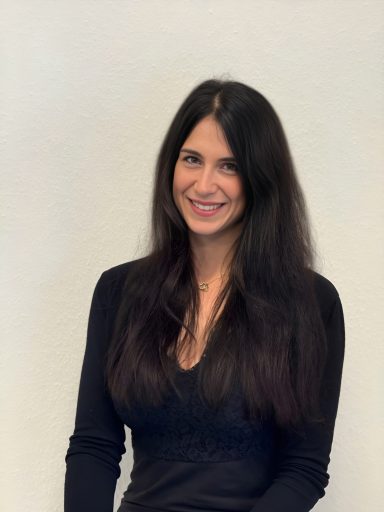 Frau mit langen, schwarzen Haaren, die lächelnd in die Kamera schaut, trägt ein schwarzes Oberteil.